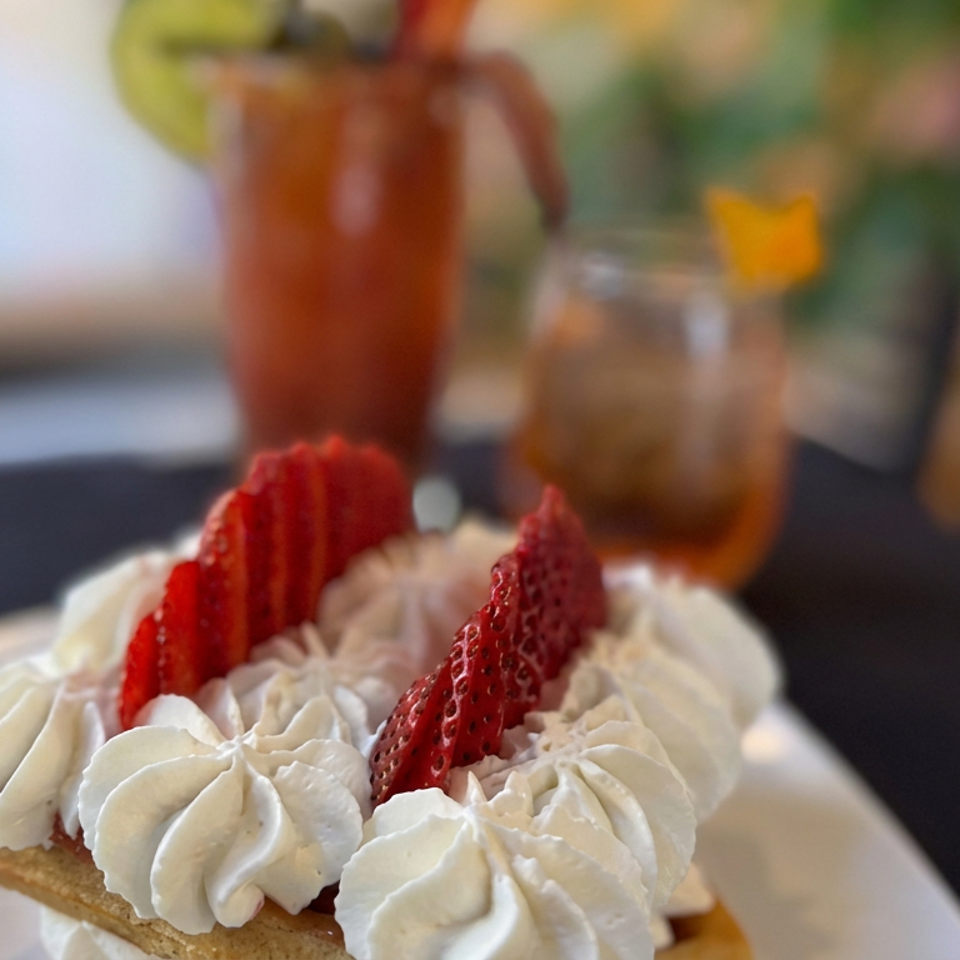 Strawberry shortcake topped with whipped cream and sliced strawberries, served on a white plate with drinks blurred in the background.