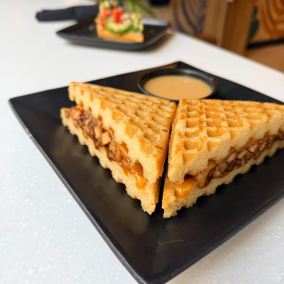 Grilled waffle sandwiches filled with melted cheese and meat, served with dipping sauce on a black plate.