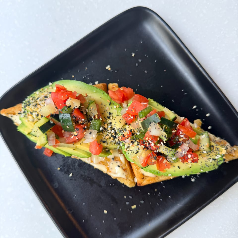 Avocado toast topped with diced tomatoes, cucumbers, onions, and mixed seeds on a black plate.