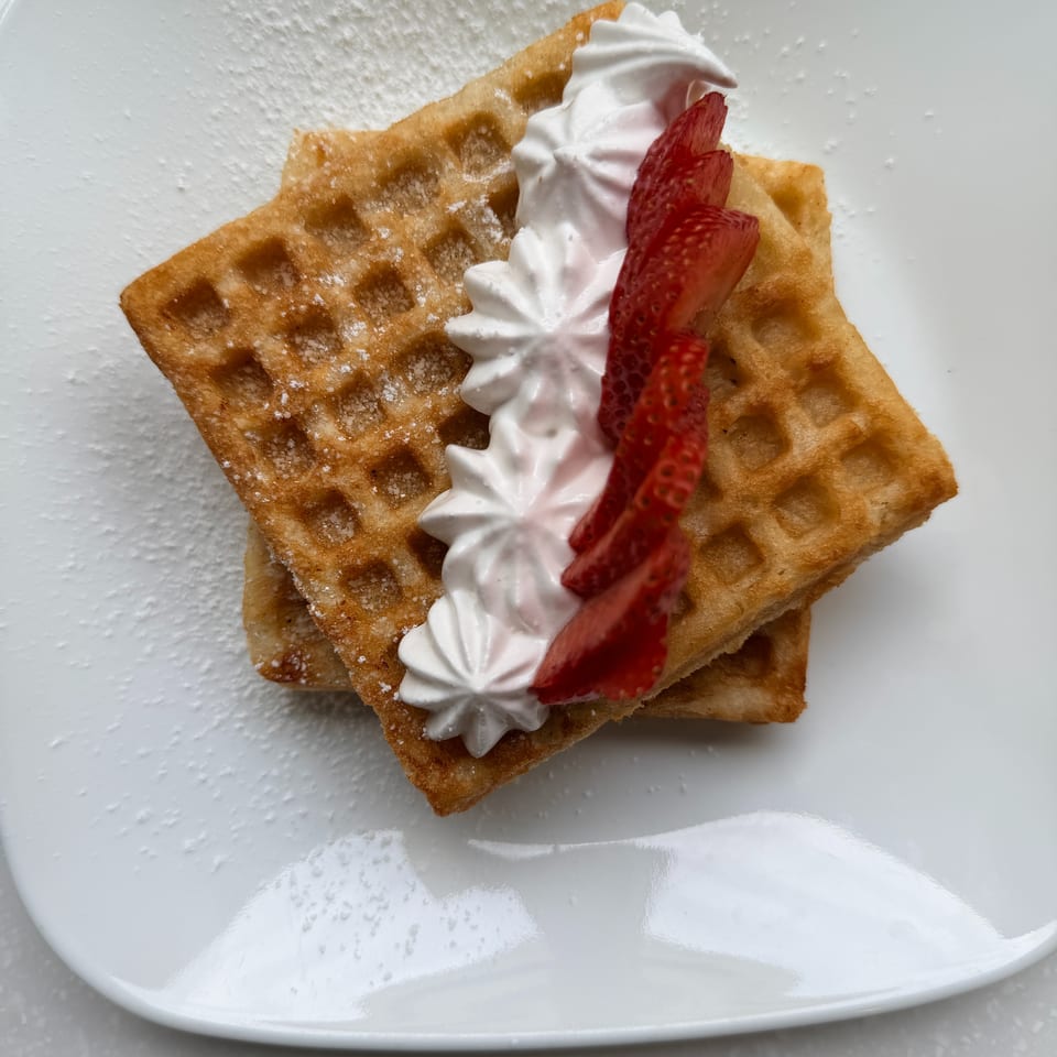 Golden waffles dusted with powdered sugar, topped with whipped cream and sliced strawberries on a white plate.