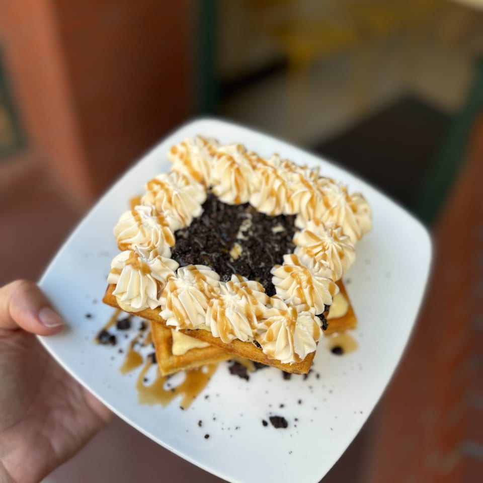 Stacked waffles topped with piped cream, caramel drizzle, and cookie crumbs on a white plate held by hand.