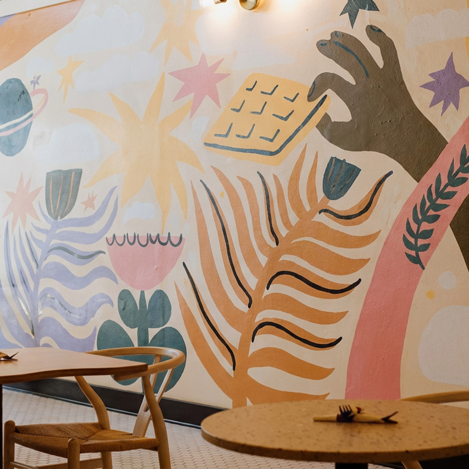 Restaurant dining area with wooden tables, woven chairs, plants, and a colorful abstract mural covering the wall.