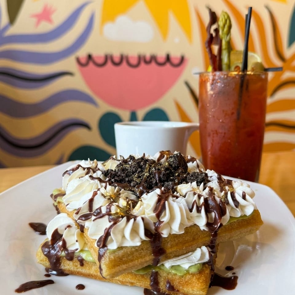 Layered waffle sandwich topped with whipped cream, chocolate drizzle, cookie crumbs, and gold sprinkles, beside a garnished red drink.